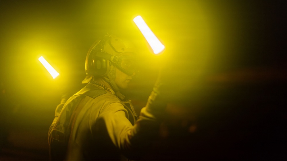 A person wearing a helmet with goggles and protective gear is holding two illuminated light wands in a dark, yellow-hued environment. The scene appears to depict a flight deck or airfield operations worker, likely a aircraft handler or director, using marshalling wands during nighttime operations. The yellow atmospheric glow and dramatic lighting create a high-contrast, visually striking composition.