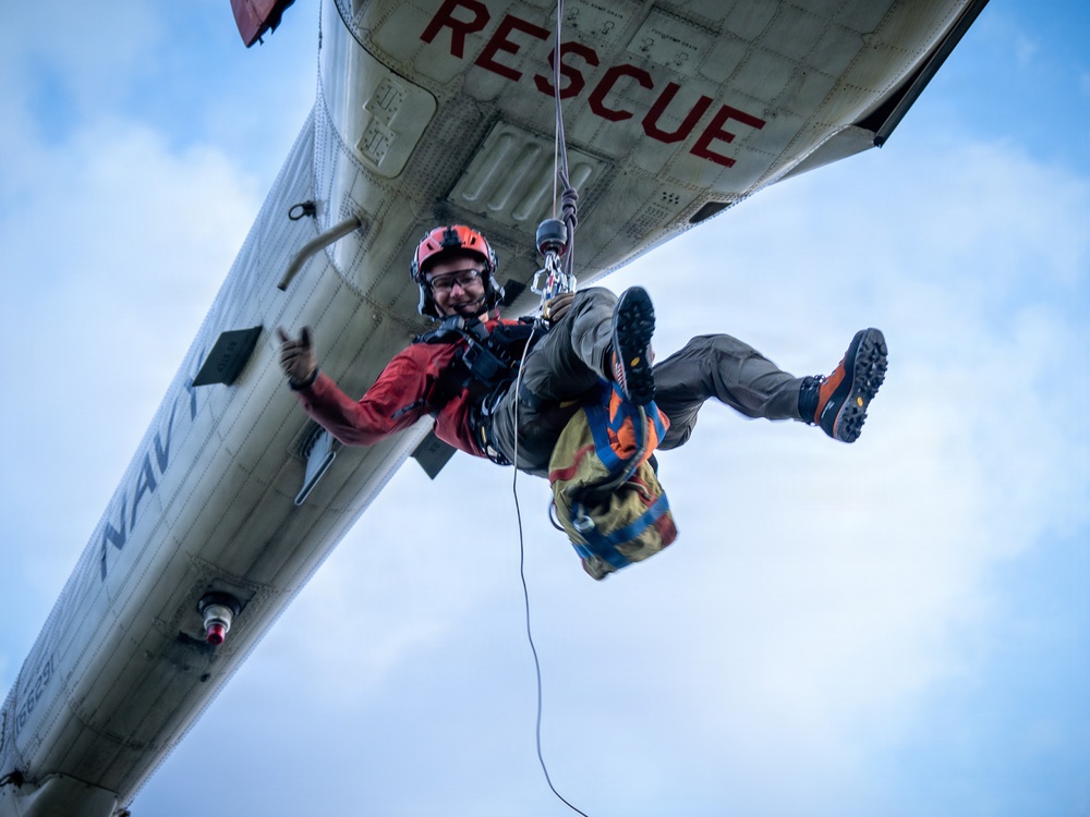 A rescue swimmer or search-and-rescue (SAR) personnel is being hoisted by a hoist cable beneath a U.S. Navy helicopter marked 'RESCUE' and 'NAVY'. The individual is wearing a red helmet, red jacket, harness, and hiking boots, and appears to be giving a thumbs-up gesture while carrying a colorful equipment bag. This appears to be either an active rescue operation or a training exercise.