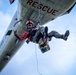 A rescue swimmer or search-and-rescue (SAR) personnel is being hoisted by a hoist cable beneath a U.S. Navy helicopter marked 'RESCUE' and 'NAVY'. The individual is wearing a red helmet, red jacket, harness, and hiking boots, and appears to be giving a thumbs-up gesture while carrying a colorful equipment bag. This appears to be either an active rescue operation or a training exercise.