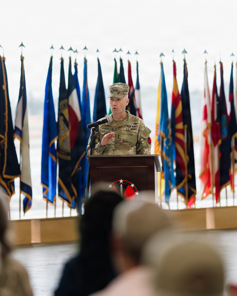 A U.S. Army officer with the name tag 'HARRINGTON' and rank insignia of a Lieutenant General (three stars) is speaking at a podium during a formal military ceremony. The podium bears a red and white insignia with stars, and the backdrop features numerous military and U.S. state flags. An audience is visible in the foreground, seated and facing the speaker.