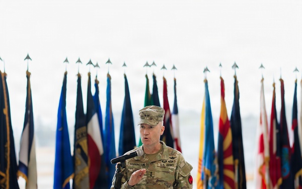 A U.S. Army officer with the name tag 'HARRINGTON' and rank insignia of a Lieutenant General (three stars) is speaking at a podium during a formal military ceremony. The podium bears a red and white insignia with stars, and the backdrop features numerous military and U.S. state flags. An audience is visible in the foreground, seated and facing the speaker.