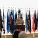 A U.S. Army officer with the name tag 'HARRINGTON' and rank insignia of a Lieutenant General (three stars) is speaking at a podium during a formal military ceremony. The podium bears a red and white insignia with stars, and the backdrop features numerous military and U.S. state flags. An audience is visible in the foreground, seated and facing the speaker.