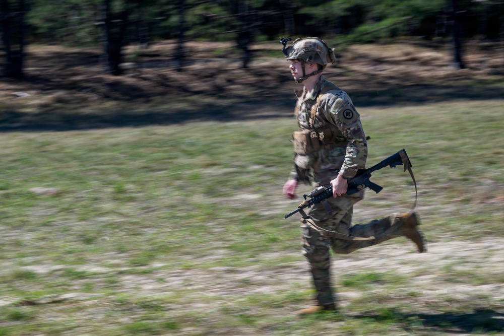 A U.S. Army soldier in full combat gear is running across an open field during what appears to be a training exercise. The soldier is wearing MultiCam (OCP) camouflage uniform, a tactical helmet with a night vision device mount, and is carrying an M4/M16-style rifle. The image uses motion blur (panning technique) to convey speed and movement.