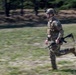 A U.S. Army soldier in full combat gear is running across an open field during what appears to be a training exercise. The soldier is wearing MultiCam (OCP) camouflage uniform, a tactical helmet with a night vision device mount, and is carrying an M4/M16-style rifle. The image uses motion blur (panning technique) to convey speed and movement.