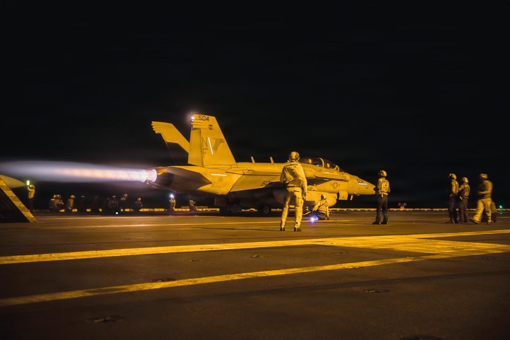 A U.S. Navy carrier-based jet aircraft, numbered 504, is preparing for a night launch on an aircraft carrier flight deck. The aircraft's afterburner is visibly ignited, producing a bright exhaust plume to the left. Multiple flight deck crew members in full gear are positioned around the aircraft observing or directing the launch sequence.