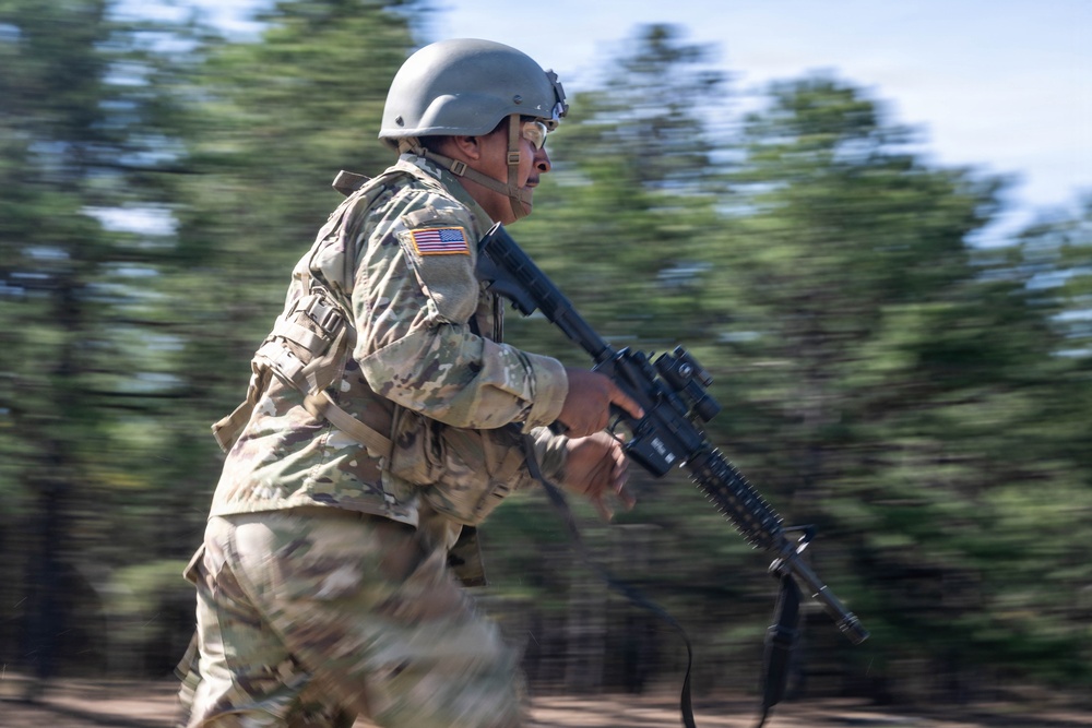 A U.S. Army soldier in full combat gear is captured mid-sprint in a dynamic panning photograph. The soldier is carrying an assault rifle and wearing MultiCam (OCP) camouflage uniform with a combat helmet and American flag patch visible on the right shoulder. The motion blur in the background suggests high speed of movement, likely during a training exercise.