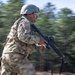 A U.S. Army soldier in full combat gear is captured mid-sprint in a dynamic panning photograph. The soldier is carrying an assault rifle and wearing MultiCam (OCP) camouflage uniform with a combat helmet and American flag patch visible on the right shoulder. The motion blur in the background suggests high speed of movement, likely during a training exercise.