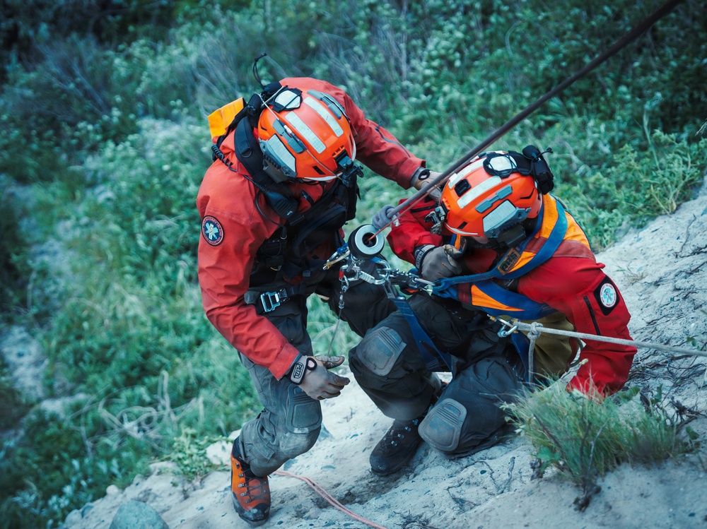 Two search and rescue personnel in orange helmets and red jackets are working together on a steep rocky slope. They appear to be managing rope and rigging equipment, likely during a technical rescue operation. Both individuals are equipped with harnesses, carabiners, and other climbing/rescue gear.