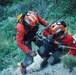 Two search and rescue personnel in orange helmets and red jackets are working together on a steep rocky slope. They appear to be managing rope and rigging equipment, likely during a technical rescue operation. Both individuals are equipped with harnesses, carabiners, and other climbing/rescue gear.