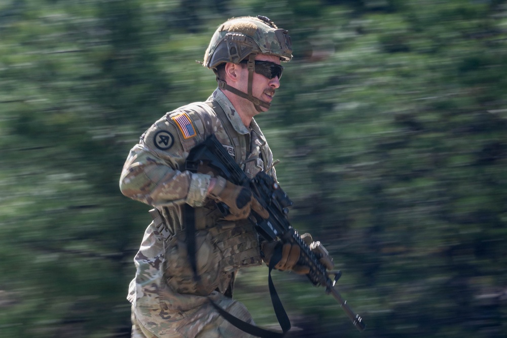 A U.S. Army soldier in full combat gear is captured mid-movement, appearing to be running or moving rapidly through a wooded area. The soldier is wearing MultiCam (OCP) camouflage uniform with a tactical helmet, ballistic eyewear, and is carrying an assault rifle. The background is heavily motion-blurred, suggesting a panning camera technique was used to capture the subject in motion.