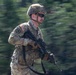 A U.S. Army soldier in full combat gear is captured mid-movement, appearing to be running or moving rapidly through a wooded area. The soldier is wearing MultiCam (OCP) camouflage uniform with a tactical helmet, ballistic eyewear, and is carrying an assault rifle. The background is heavily motion-blurred, suggesting a panning camera technique was used to capture the subject in motion.