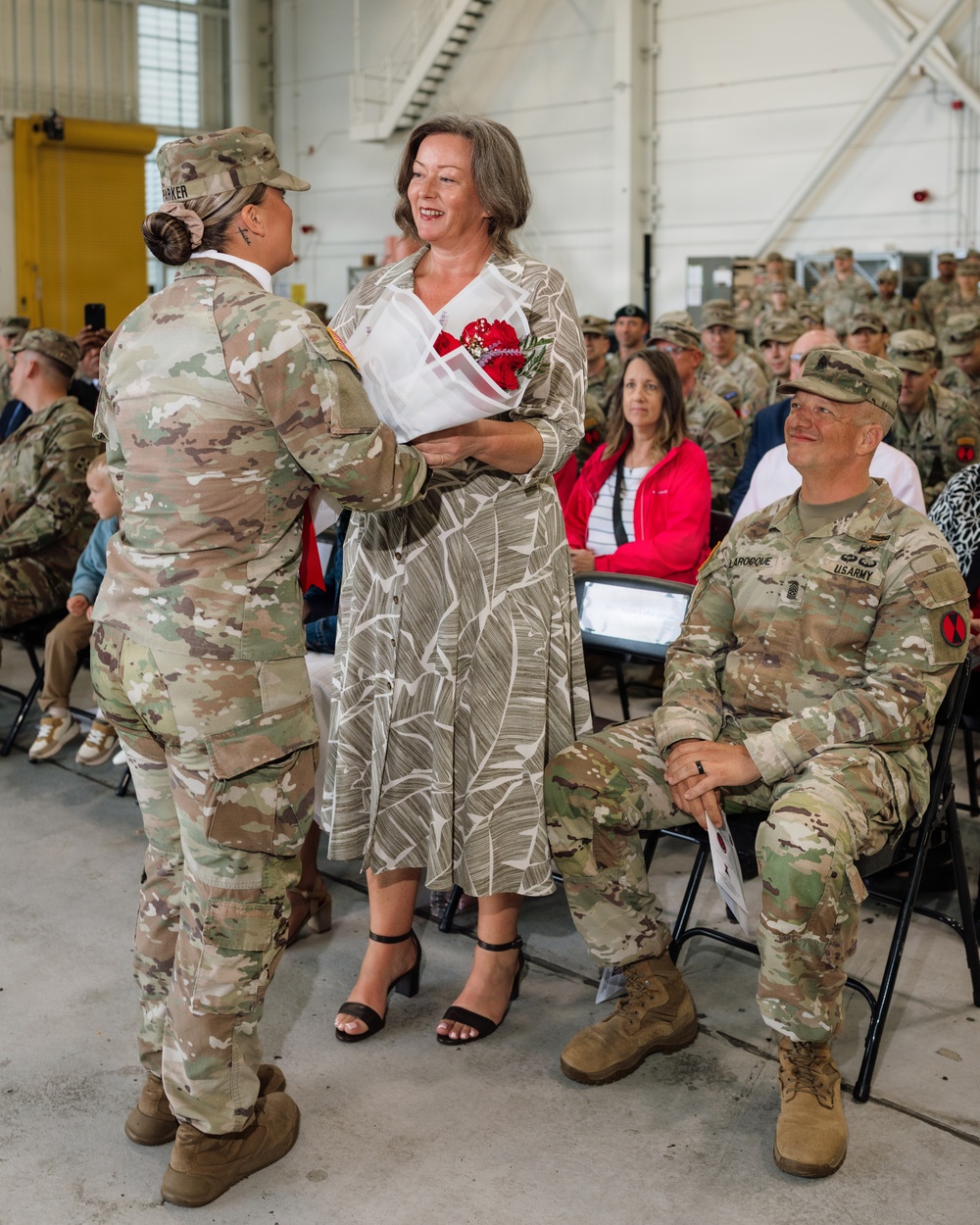 A military ceremony taking place inside what appears to be a hangar or large military facility. A female soldier in OCP (Operational Camouflage Pattern) uniform is presenting red flowers to a civilian woman in a floral dress, while a seated male U.S. Army soldier (name tape reads 'LAROCQUE') looks on smiling. Rows of uniformed soldiers and some civilians are seated in the background as an audience.