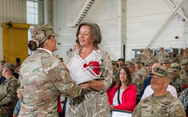 A military ceremony taking place inside what appears to be a hangar or large military facility. A female soldier in OCP (Operational Camouflage Pattern) uniform is presenting red flowers to a civilian woman in a floral dress, while a seated male U.S. Army soldier (name tape reads 'LAROCQUE') looks on smiling. Rows of uniformed soldiers and some civilians are seated in the background as an audience.