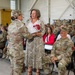 A military ceremony taking place inside what appears to be a hangar or large military facility. A female soldier in OCP (Operational Camouflage Pattern) uniform is presenting red flowers to a civilian woman in a floral dress, while a seated male U.S. Army soldier (name tape reads 'LAROCQUE') looks on smiling. Rows of uniformed soldiers and some civilians are seated in the background as an audience.