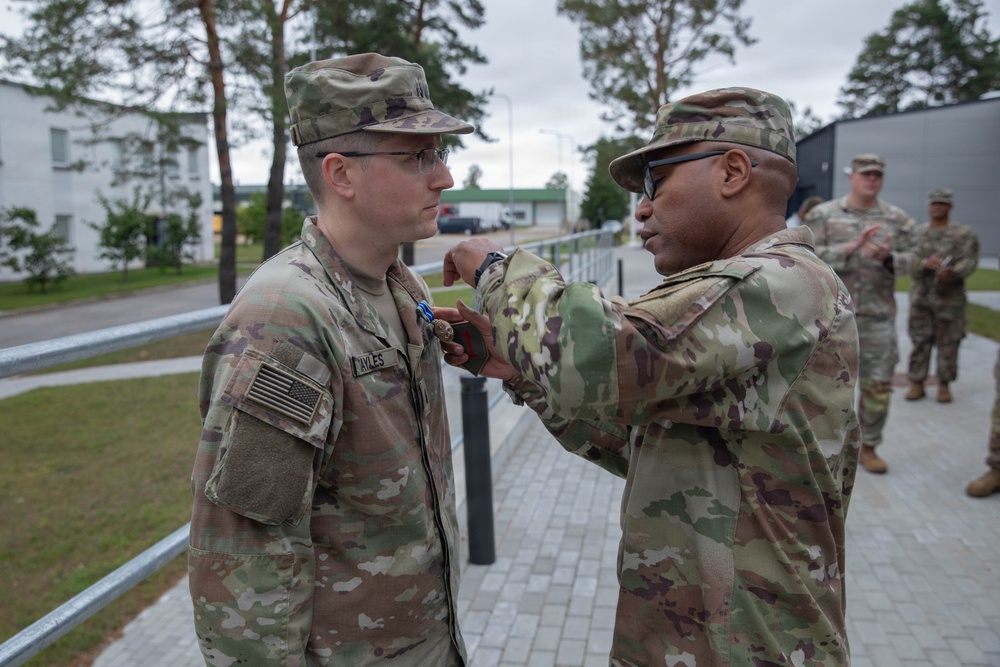 A U.S. Army medal pinning ceremony is taking place outdoors at what appears to be a military installation. A senior soldier is pinning a medal onto the chest of a soldier identified by the name tape 'MYLES', while other soldiers observe in the background. Both primary subjects are wearing OCP (Operational Camouflage Pattern) uniforms with U.S. flag patches.