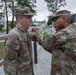 A U.S. Army medal pinning ceremony is taking place outdoors at what appears to be a military installation. A senior soldier is pinning a medal onto the chest of a soldier identified by the name tape 'MYLES', while other soldiers observe in the background. Both primary subjects are wearing OCP (Operational Camouflage Pattern) uniforms with U.S. flag patches.