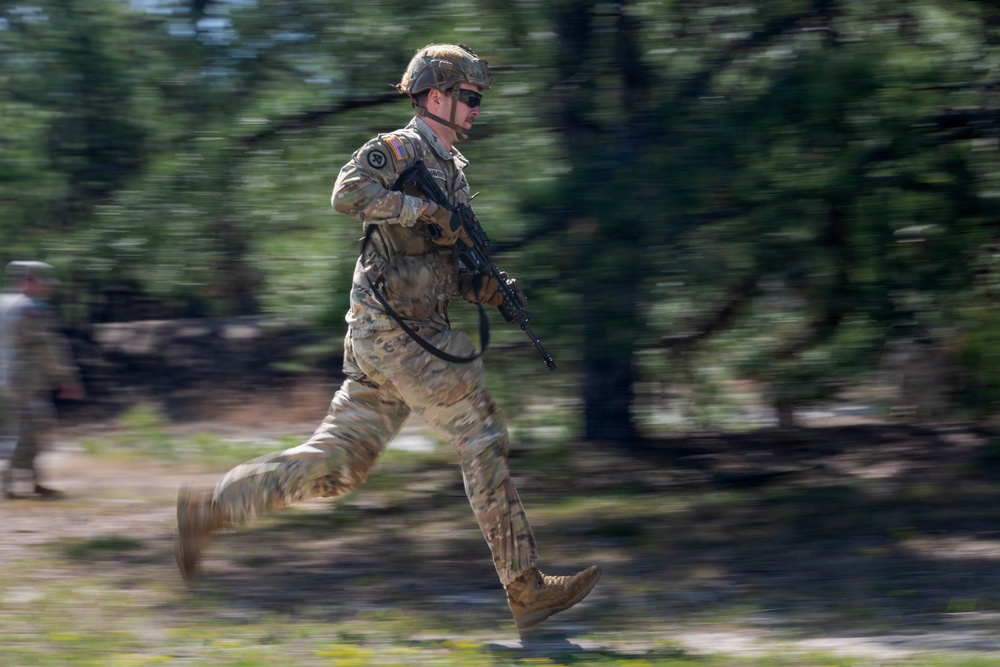 A U.S. Army soldier in full combat gear is captured mid-sprint in what appears to be a training exercise. The soldier is wearing MultiCam (OCP) camouflage, a tactical helmet, and sunglasses while carrying an assault rifle. The motion-blur background technique used by the photographer emphasizes the speed and dynamic movement of the subject.