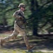 A U.S. Army soldier in full combat gear is captured mid-sprint in what appears to be a training exercise. The soldier is wearing MultiCam (OCP) camouflage, a tactical helmet, and sunglasses while carrying an assault rifle. The motion-blur background technique used by the photographer emphasizes the speed and dynamic movement of the subject.