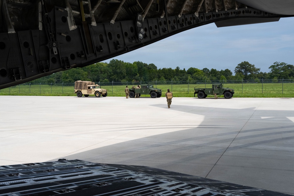 The image is taken from inside the rear cargo bay of a large military transport aircraft, looking out through the open ramp onto a tarmac. Three military Humvees (HMMWVs) are staged on the flight line, with several soldiers in camouflage uniforms moving around them, apparently preparing for loading operations. The setting appears to be a military airfield surrounded by green trees under a partly cloudy sky.