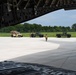 The image is taken from inside the rear cargo bay of a large military transport aircraft, looking out through the open ramp onto a tarmac. Three military Humvees (HMMWVs) are staged on the flight line, with several soldiers in camouflage uniforms moving around them, apparently preparing for loading operations. The setting appears to be a military airfield surrounded by green trees under a partly cloudy sky.