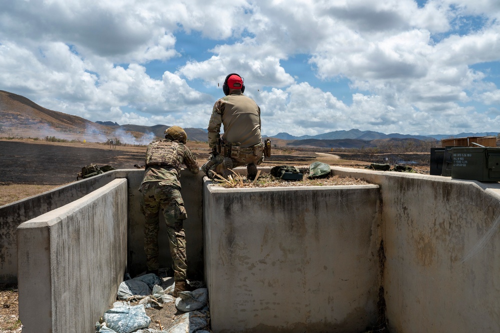 Two military personnel are positioned behind a concrete firing position or bunker, observing a downrange area with visible smoke in the distance. The scene appears to be a live-fire training exercise on a military range, with mountainous terrain in the background. One individual wears full camouflage with tactical gear, while the other wears tan/khaki clothing with a red hearing protection headset.