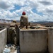 Two military personnel are positioned behind a concrete firing position or bunker, observing a downrange area with visible smoke in the distance. The scene appears to be a live-fire training exercise on a military range, with mountainous terrain in the background. One individual wears full camouflage with tactical gear, while the other wears tan/khaki clothing with a red hearing protection headset.