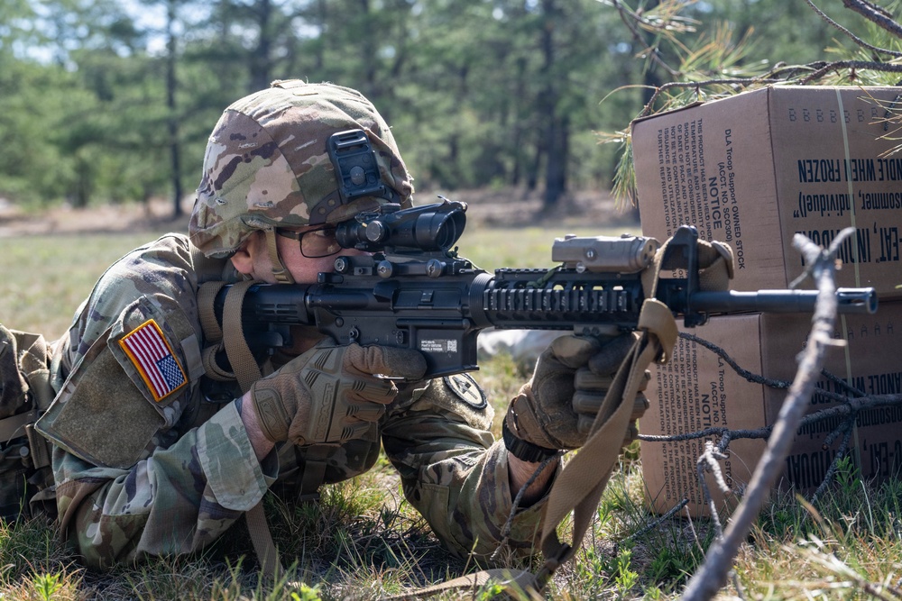 A U.S. Army soldier in MultiCam/OCP camouflage uniform is in a prone firing position, aiming an M4 carbine rifle equipped with an optic sight. The soldier is using cardboard boxes marked with DLA (Defense Logistics Agency) 'Troop Support' labeling as cover. The setting appears to be a wooded training area, consistent with a military field exercise.