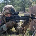 A U.S. Army soldier in MultiCam/OCP camouflage uniform is in a prone firing position, aiming an M4 carbine rifle equipped with an optic sight. The soldier is using cardboard boxes marked with DLA (Defense Logistics Agency) 'Troop Support' labeling as cover. The setting appears to be a wooded training area, consistent with a military field exercise.