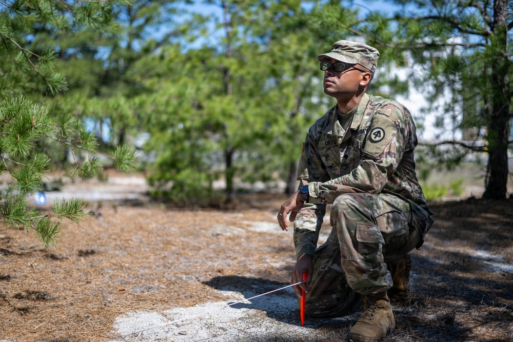 A U.S. Army soldier identified by the name tape 'ORTIZ' is crouching in a wooded, sandy training area while looking upward and to the side. The soldier is wearing Operational Camouflage Pattern (OCP) uniform with an Army patch on the left shoulder and sunglasses. A red marker stake with a white cord or string is visible near the ground in front of the soldier.