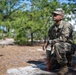 A U.S. Army soldier identified by the name tape 'ORTIZ' is crouching in a wooded, sandy training area while looking upward and to the side. The soldier is wearing Operational Camouflage Pattern (OCP) uniform with an Army patch on the left shoulder and sunglasses. A red marker stake with a white cord or string is visible near the ground in front of the soldier.