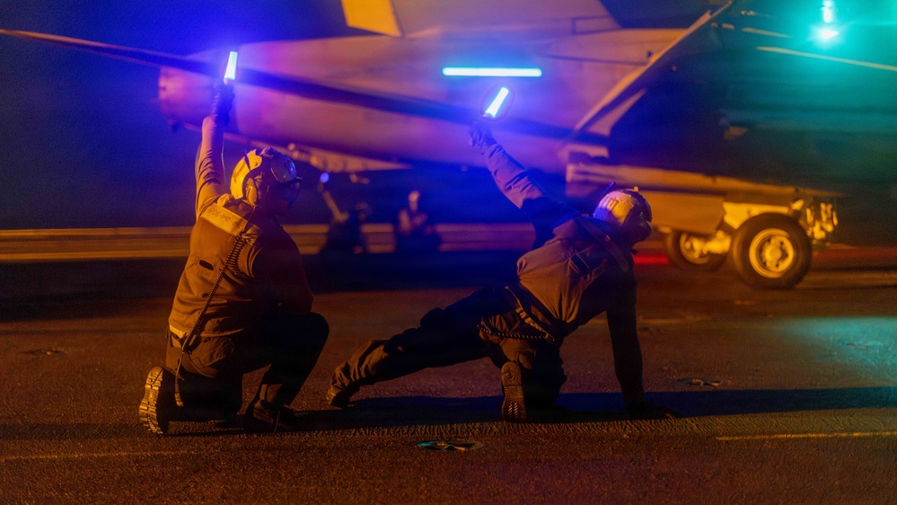 Two military flight deck personnel are conducting night operations on what appears to be an aircraft carrier flight deck. They are using illuminated wands/batons to guide or signal an aircraft during low-light conditions. The scene is lit by a combination of blue aircraft lighting and ambient orange/teal light sources.