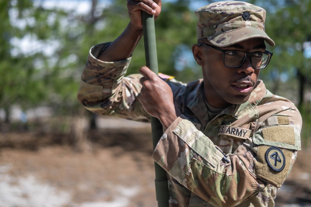 A U.S. Army soldier in Operational Camouflage Pattern (OCP) uniform is handling a green metal pole or rod outdoors. The soldier is wearing glasses and a patrol cap, with a unit patch visible on the left sleeve. The background shows a wooded or scrubland environment with blurred trees and sandy ground.