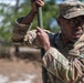 A U.S. Army soldier in Operational Camouflage Pattern (OCP) uniform is handling a green metal pole or rod outdoors. The soldier is wearing glasses and a patrol cap, with a unit patch visible on the left sleeve. The background shows a wooded or scrubland environment with blurred trees and sandy ground.