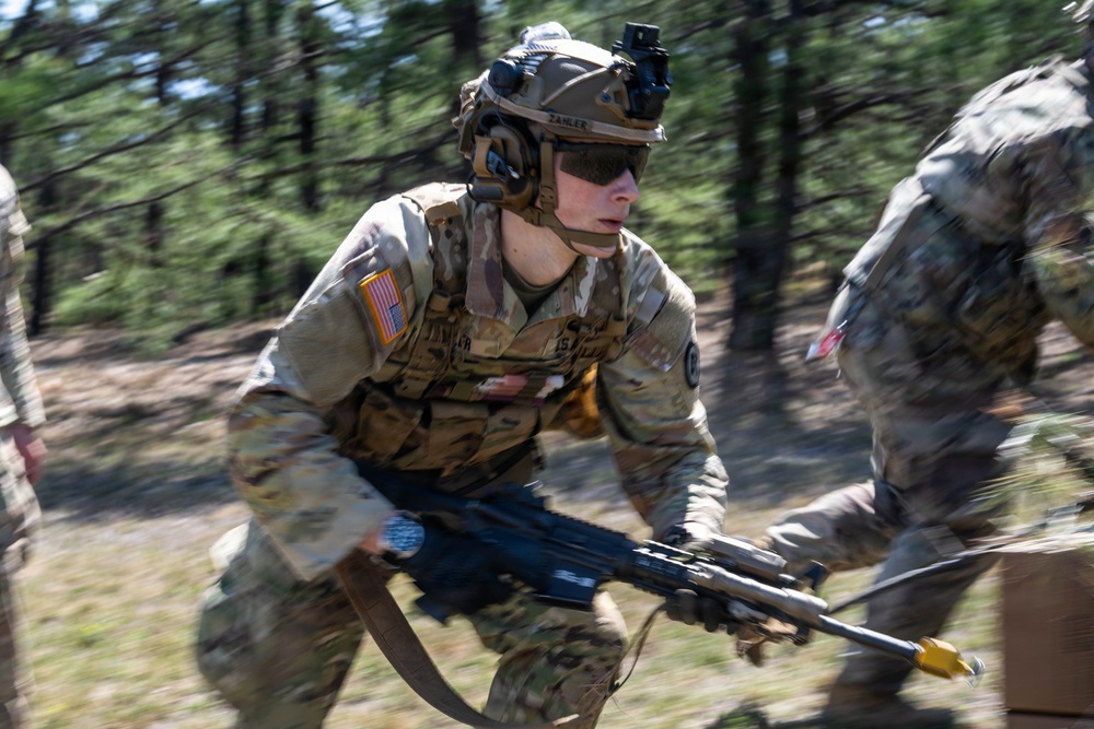 A U.S. Army soldier in full combat gear is captured mid-movement in a wooded training environment, appearing to be conducting a field exercise. The soldier is wearing MultiCam (OCP) camouflage, a tactical helmet with night vision mount, and is carrying what appears to be an M249 SAW or similar light machine gun. Motion blur in the image suggests rapid movement during what appears to be a military training exercise.
