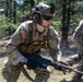 A U.S. Army soldier in full combat gear is captured mid-movement in a wooded training environment, appearing to be conducting a field exercise. The soldier is wearing MultiCam (OCP) camouflage, a tactical helmet with night vision mount, and is carrying what appears to be an M249 SAW or similar light machine gun. Motion blur in the image suggests rapid movement during what appears to be a military training exercise.
