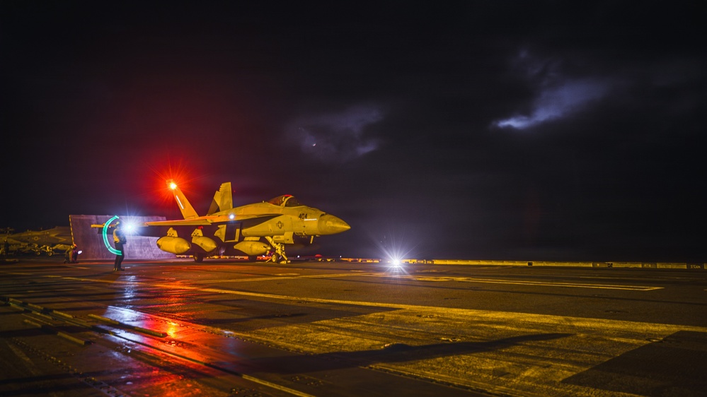 A military jet aircraft, appearing to be an F/A-18 Hornet or Super Hornet, is positioned on a wet aircraft carrier flight deck at night. The scene is illuminated by red and white deck lights, with a stormy, lightning-lit sky visible in the background. Ground crew personnel are visible near the aircraft, suggesting pre-launch or post-landing operations.