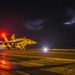 A military jet aircraft, appearing to be an F/A-18 Hornet or Super Hornet, is positioned on a wet aircraft carrier flight deck at night. The scene is illuminated by red and white deck lights, with a stormy, lightning-lit sky visible in the background. Ground crew personnel are visible near the aircraft, suggesting pre-launch or post-landing operations.
