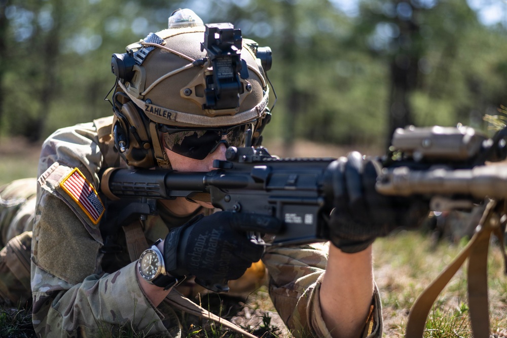 A U.S. Army soldier identified as 'Zahler' is in a prone firing position aiming a rifle with an attached optic and accessories. The soldier is wearing full tactical gear including a FAST-style helmet with night vision device mount, multicam uniform, tactical gloves, and a wristwatch. The setting appears to be an outdoor training range or field exercise environment.