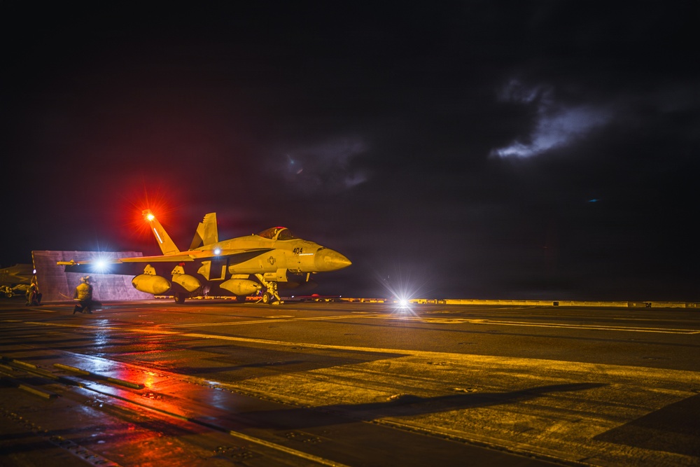 A military jet aircraft, marked with the number '404', is positioned on a wet aircraft carrier flight deck at night. The scene is illuminated by red and white deck lights reflecting off the rain-soaked surface, with a stormy, cloud-filled sky in the background. At least one deck crew member is visible near the aircraft, suggesting pre-launch or post-landing operations.