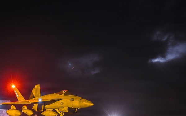 A military jet aircraft, marked with the number '404', is positioned on a wet aircraft carrier flight deck at night. The scene is illuminated by red and white deck lights reflecting off the rain-soaked surface, with a stormy, cloud-filled sky in the background. At least one deck crew member is visible near the aircraft, suggesting pre-launch or post-landing operations.