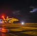 A military jet aircraft, marked with the number '404', is positioned on a wet aircraft carrier flight deck at night. The scene is illuminated by red and white deck lights reflecting off the rain-soaked surface, with a stormy, cloud-filled sky in the background. At least one deck crew member is visible near the aircraft, suggesting pre-launch or post-landing operations.