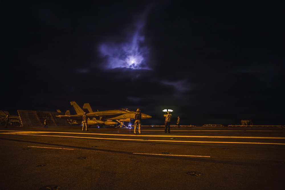 A nighttime scene on what appears to be an aircraft carrier flight deck, with a military jet aircraft (likely an F/A-18 Hornet or Super Hornet) positioned on the deck. Several deck crew personnel are visible, one holding a wand light, under a dramatic sky illuminated by moonlight breaking through clouds. The scene is lit primarily by amber deck lighting and the crew member's handheld light.