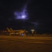 A nighttime scene on what appears to be an aircraft carrier flight deck, with a military jet aircraft (likely an F/A-18 Hornet or Super Hornet) positioned on the deck. Several deck crew personnel are visible, one holding a wand light, under a dramatic sky illuminated by moonlight breaking through clouds. The scene is lit primarily by amber deck lighting and the crew member's handheld light.