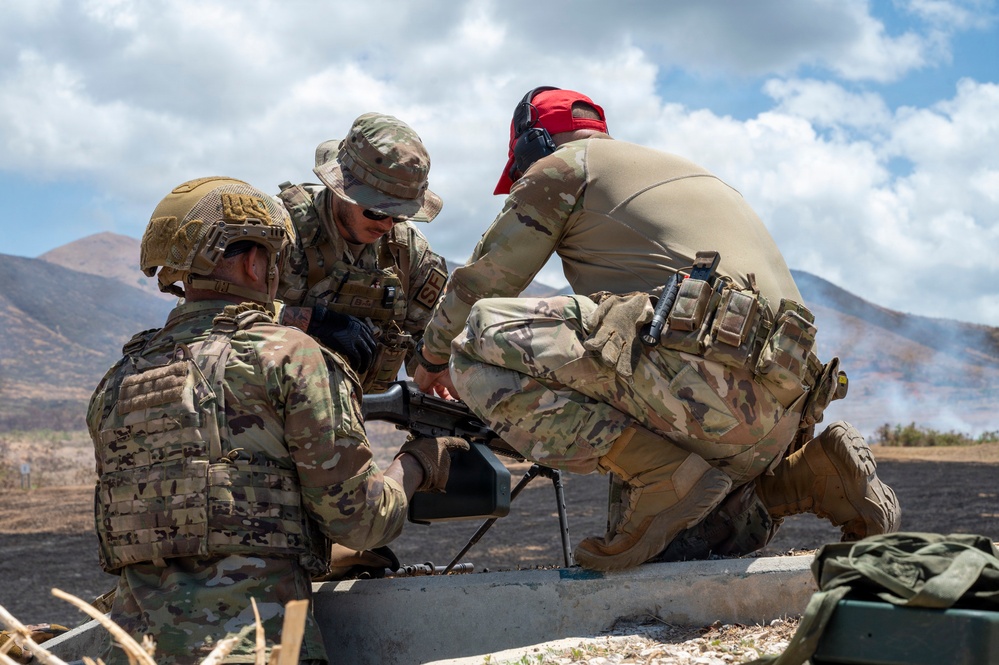Three military personnel in multicam/OCP camouflage uniforms are gathered around a weapon system, appearing to conduct training or maintenance at an outdoor range. The setting appears to be an arid, mountainous training area with smoke visible in the background. One individual wears a red cap with hearing protection, another wears a boonie hat, and the third wears a tactical helmet.