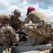 Three military personnel in multicam/OCP camouflage uniforms are gathered around a weapon system, appearing to conduct training or maintenance at an outdoor range. The setting appears to be an arid, mountainous training area with smoke visible in the background. One individual wears a red cap with hearing protection, another wears a boonie hat, and the third wears a tactical helmet.