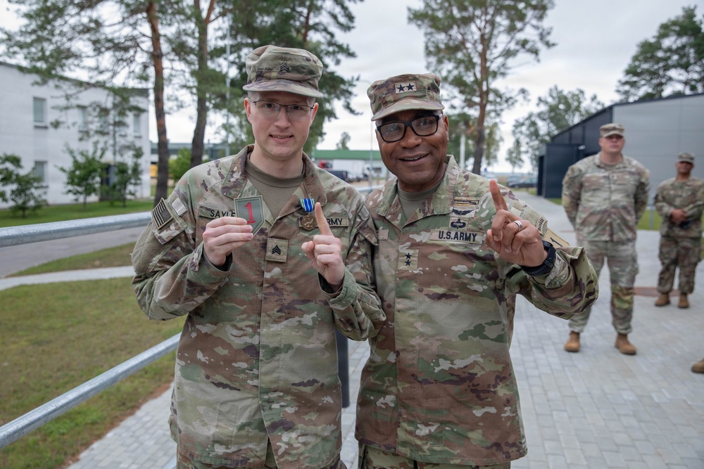Two U.S. Army soldiers pose together outdoors at what appears to be a military installation, with the soldier on the left holding up a patch and a medal. The soldier on the right wears two-star Major General rank insignia on his cap and 'U.S. ARMY' tape on his uniform. Both are making a 'number one' gesture with their index fingers, suggesting a celebratory or award-related occasion.