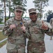 Two U.S. Army soldiers pose together outdoors at what appears to be a military installation, with the soldier on the left holding up a patch and a medal. The soldier on the right wears two-star Major General rank insignia on his cap and 'U.S. ARMY' tape on his uniform. Both are making a 'number one' gesture with their index fingers, suggesting a celebratory or award-related occasion.