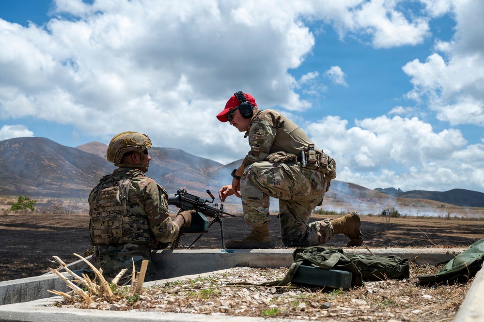 Two military personnel in MultiCam/OCP camouflage uniforms are positioned at what appears to be a firing range, with one seated behind a machine gun and the other kneeling beside them in an instructional or supervisory posture. The background shows scorched terrain with smoke visible, rolling hills, and a partly cloudy sky. The setting suggests an active live-fire training exercise.