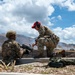 Two military personnel in MultiCam/OCP camouflage uniforms are positioned at what appears to be a firing range, with one seated behind a machine gun and the other kneeling beside them in an instructional or supervisory posture. The background shows scorched terrain with smoke visible, rolling hills, and a partly cloudy sky. The setting suggests an active live-fire training exercise.