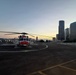 A red and white helicopter, consistent with U.S. Coast Guard livery, is parked on a rooftop helipad with green perimeter lights illuminated. The scene is captured during twilight/dusk with a city skyline visible in the background, featuring tall glass skyscrapers consistent with Seattle, Washington. The helipad appears to be atop a building, with a parking structure visible in the foreground.